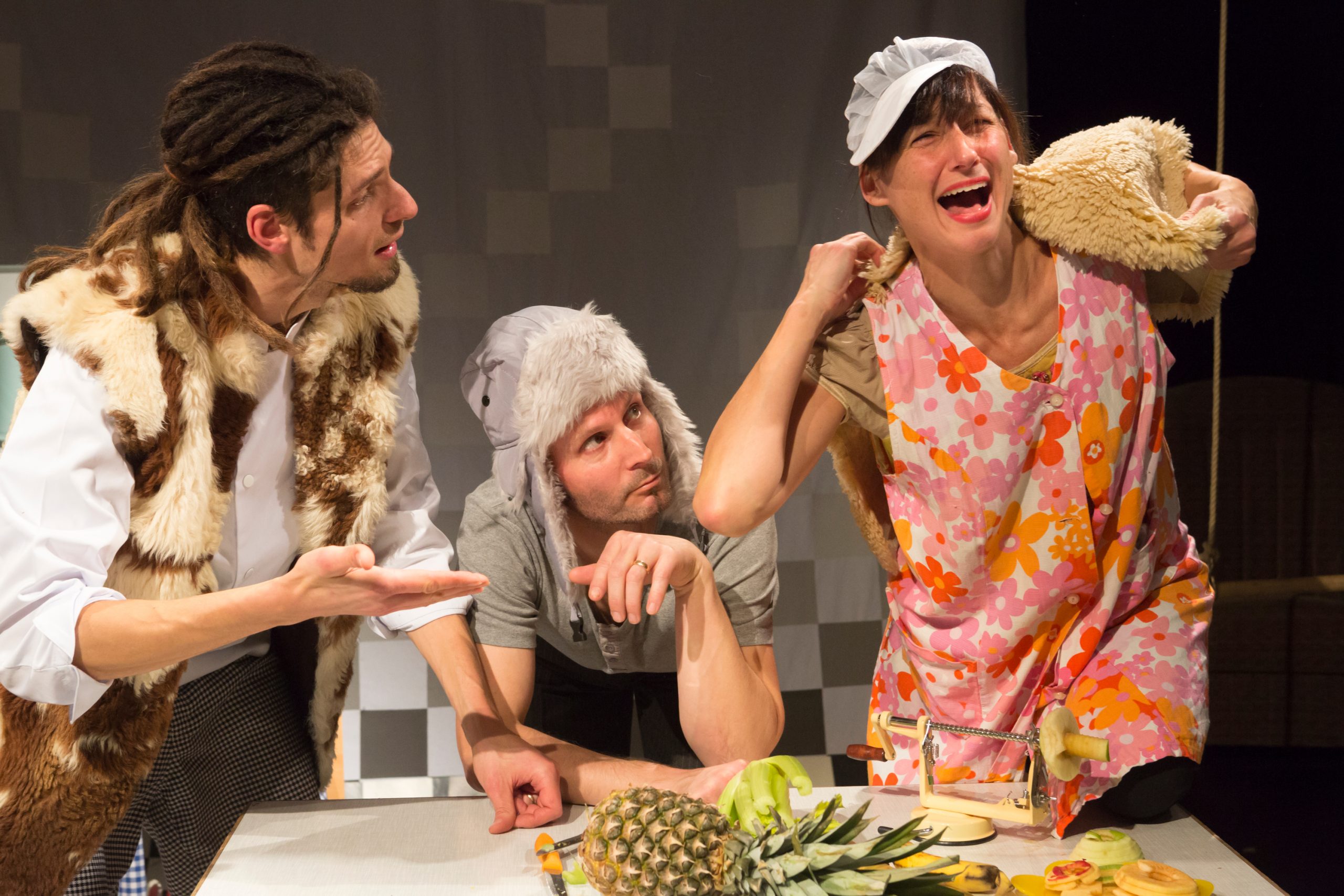 Drei Schauspieler in Kostümen bei einer Theaterszene mit einem Tisch, auf dem eine Ananas und andere Früchte liegen, einer trägt ein buntes Kleid und hält ein Stoffteil an die Schulter