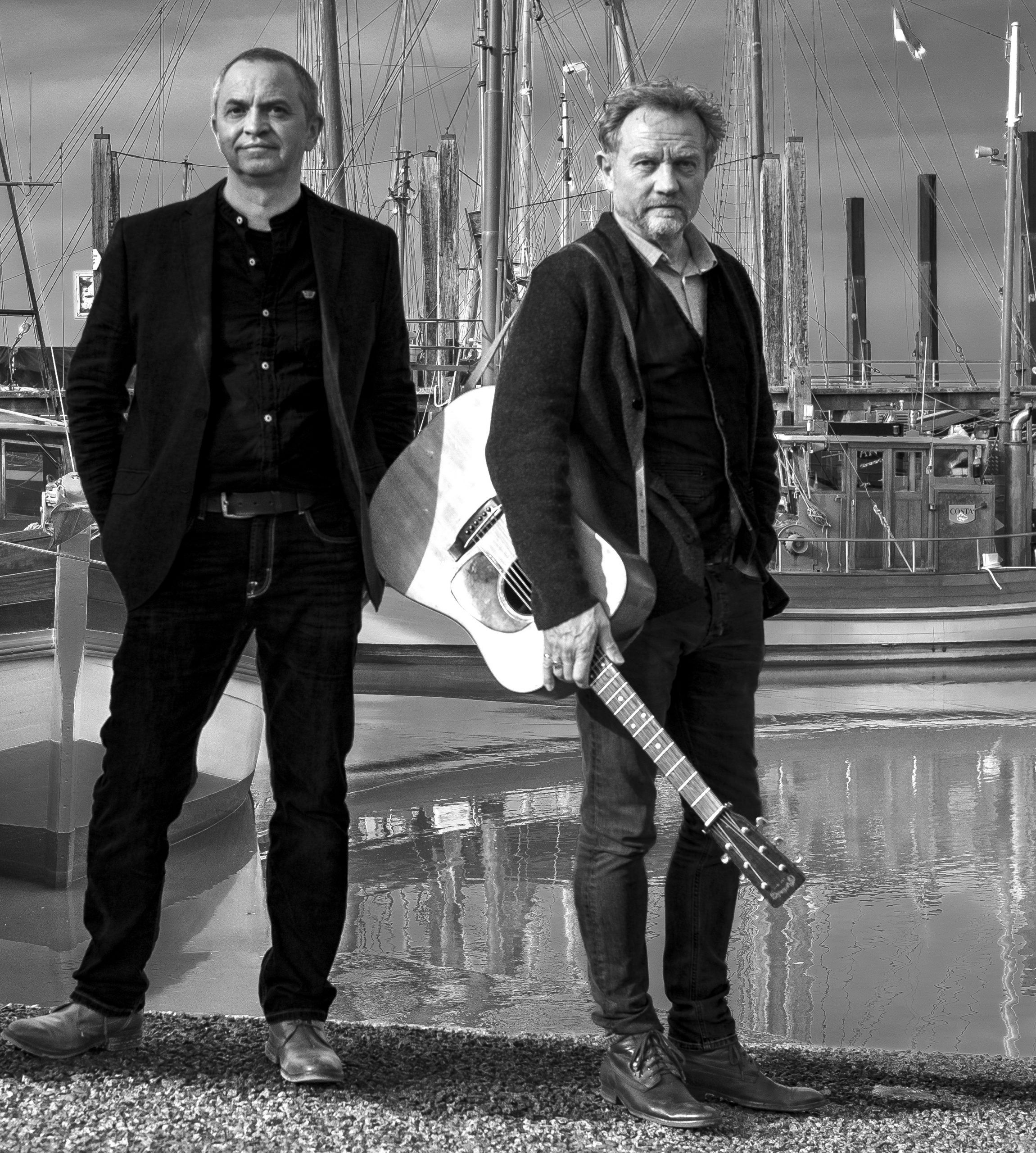Zwei Musiker stehen an einem Hafen, einer hält eine Gitarre in der Hand