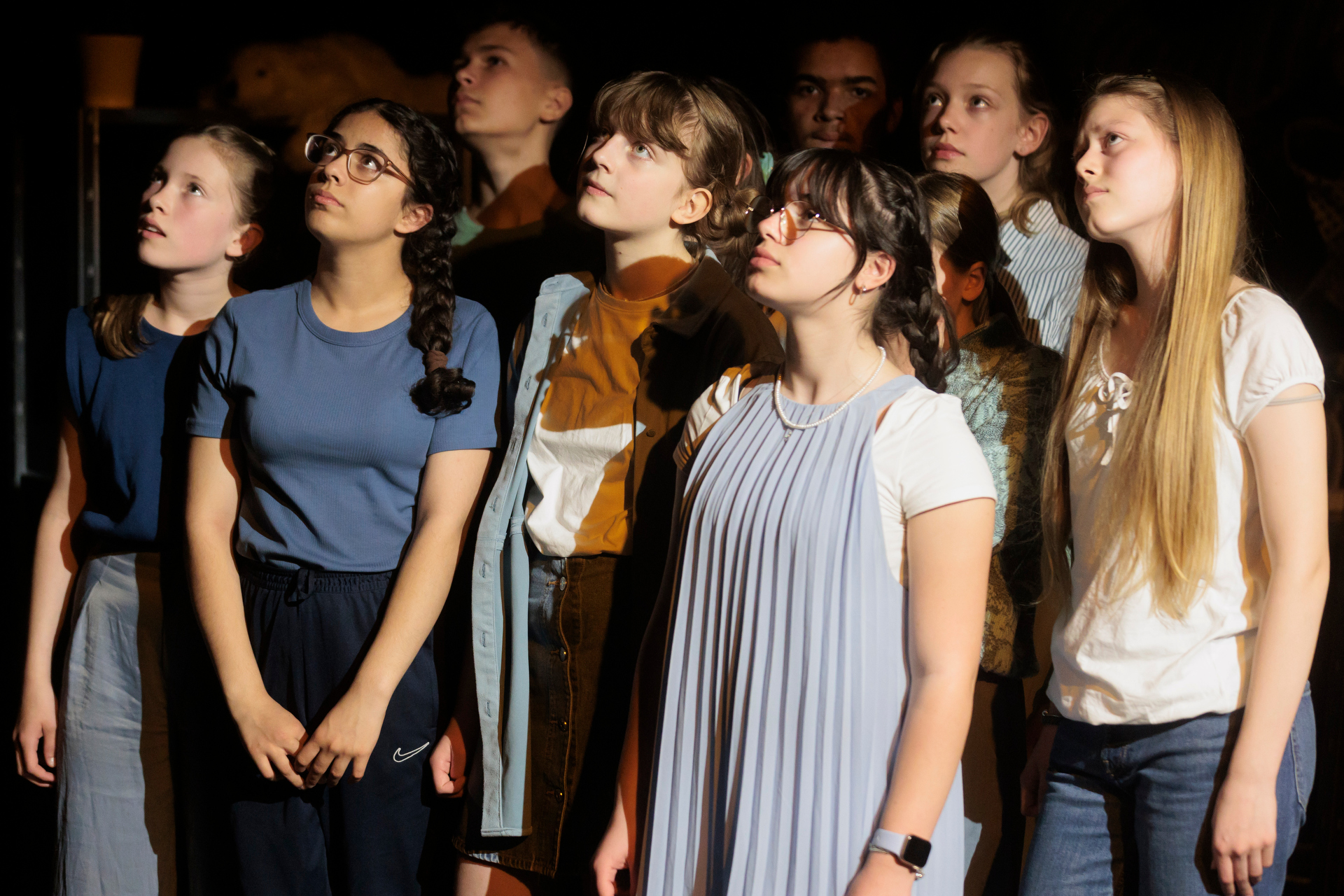 Gruppe von Jugendlichen steht eng nebeneinander auf einer Bühne, einige tragen Freizeitkleidung wie T-Shirts und Kleider, im Hintergrund dunkle Theaterkulisse